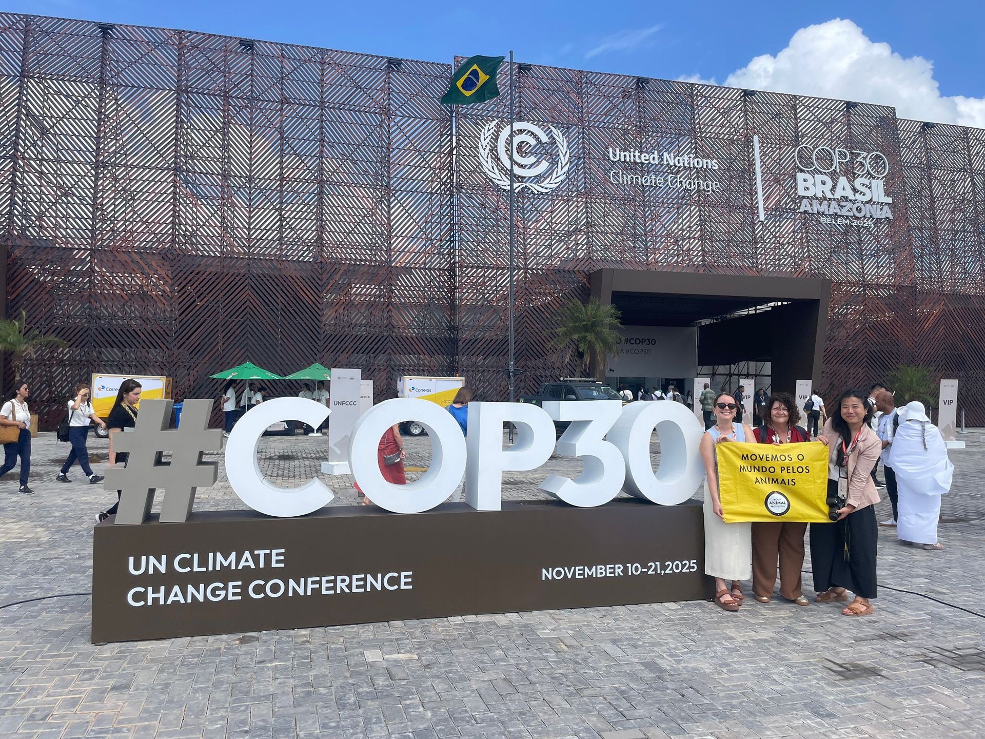 Miembros de World Animal Protection junto al gran letrero de #COP30 frente al recinto de la conferencia climática en Belém, sosteniendo una pancarta amarilla.