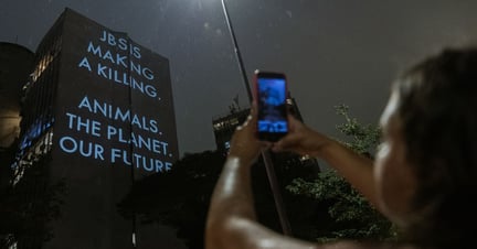 Mujer tomando una foto con su teléfono de un gran edificio con las palabras "JBS está obteniendo grandes ganancias. Animales. El planeta. Nuestro futuro." iluminadas