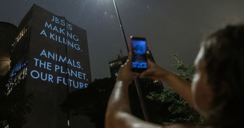 Mujer tomando una foto con su teléfono de un gran edificio con las palabras "JBS está obteniendo grandes ganancias. Animales. El planeta. Nuestro futuro." iluminadas